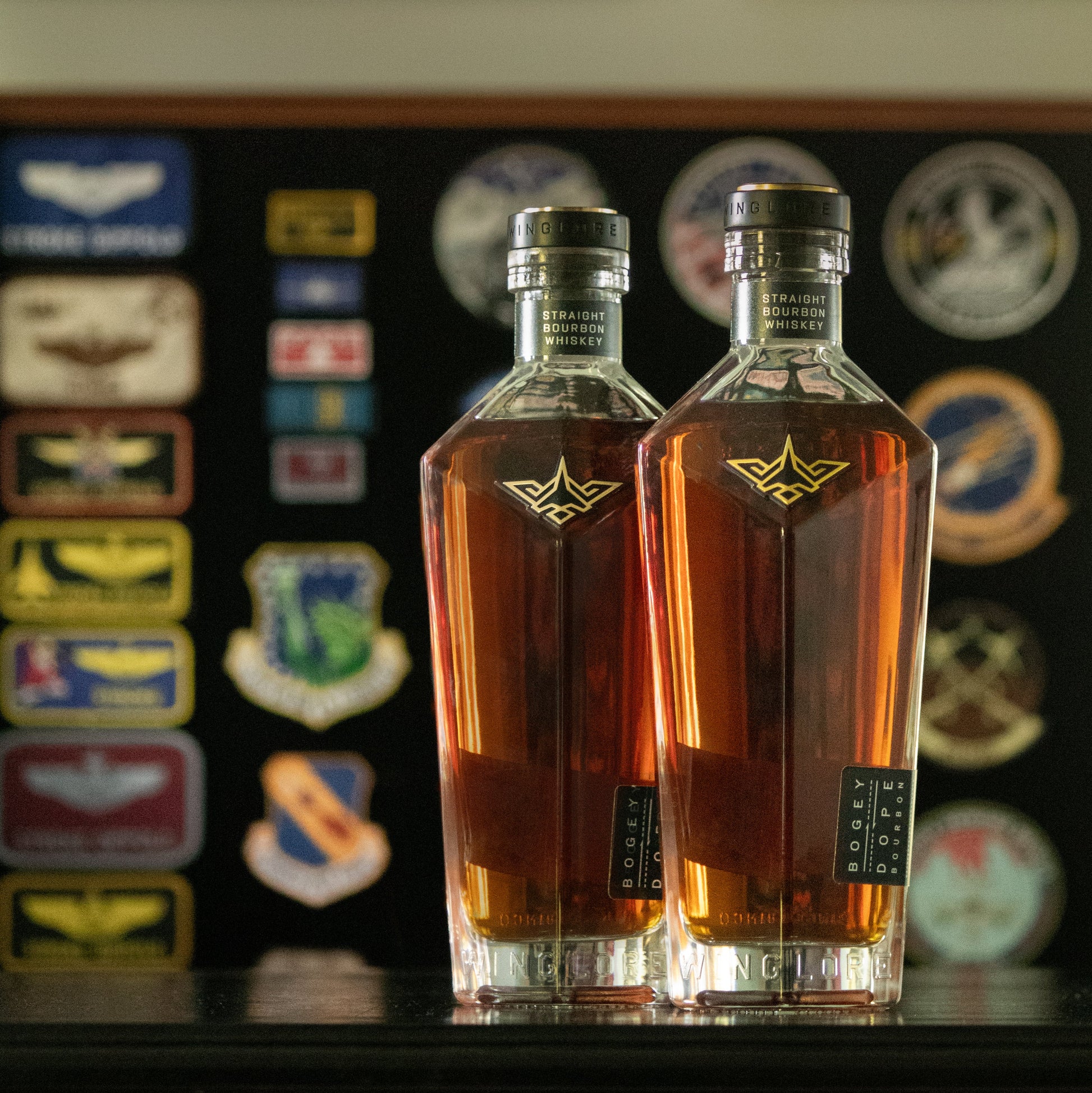 


Two clear glass bottles filled with amber liquor, standing side by side on a dark surface in front of a wall covered with military insignia and patches.